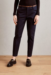 Marineblaue Hose mit taillierter Passform, ausgestattet mit einem braunen Ledergürtel, Seitentaschen und einem glatten Stoff. Schwarze Slipper mit goldenen Akzenten.