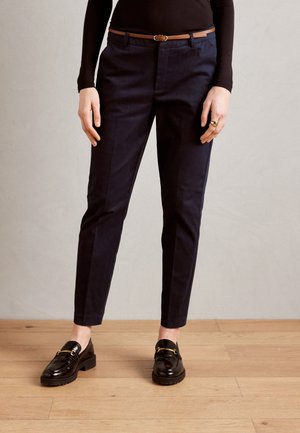 Pantalon marine à coupe ajustée, avec une ceinture en cuir marron, des poches latérales et un tissu lisse. Mocassins noirs avec des accents dorés.