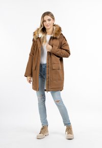 Bruine parka jas met een met bont gevoerde capuchon, voorzakken en een ritssluiting. Gedragen over een witte top en lichtblauwe ripped jeans, gecombineerd met beige laarzen.