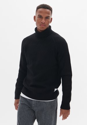 Schwarzer gerippter Rollkragenpullover mit langen Ärmeln, der eine enganliegende Passform und strukturierten Stoff aufweist. Über einem weißen T-Shirt getragen und mit grauen Jeans kombiniert.