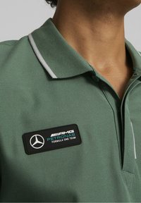 Grön polo-tröja med en texturerad tyg, som har ett svart och blått AMG Petronas Formula One Team-märke och vita krage-accenter.