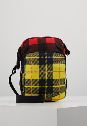 Mała torba na ramię Nike z czerwono-czarnym kratowanym wierzchem i żółto-czarnoczerwoną kratowaną przednią kieszenią, wyposażona w czarny regulowany pasek.