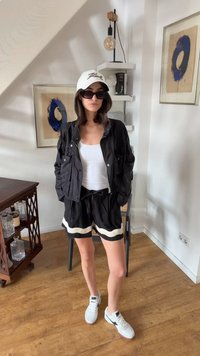Mujer con gafas de sol, gorra blanca, chaqueta negra, camiseta sin mangas blanca, pantalones cortos negros y zapatillas blancas de pie en una habitación con arte enmarcado y suelo de madera.