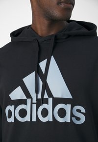 Sudadera negra con capucha y cordones, con un gran logo y marca de Adidas en azul claro en la parte delantera. Tejido texturizado.