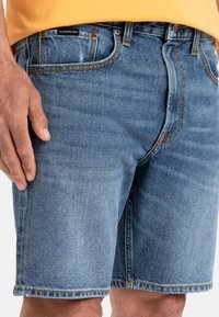 Shorts de mezclilla en azul claro, con costuras en naranja de contraste. Presenta cinco bolsillos y un corte relajado, mostrando el dobladillo justo por encima de la rodilla.
