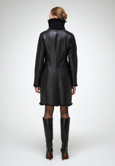 Donna che indossa un cappotto in pelle nera lungo fino al ginocchio con bordi in pelliccia, abbinato a collant neri e stivali neri con tacco al ginocchio, in piedi di spalle.