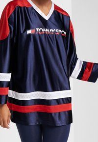 Camiseta deportiva azul marino con detalles en malla roja, rayas blancas y cuello en V. Presenta el logo "TOMMY SPORT" en blanco en el pecho.