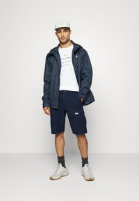 Shorts cargo navy, t-shirt gris clair, veste sombre et casquette vert menthe. Chaussures grises avec une semelle beige. Tenue décontractée pour l'extérieur.