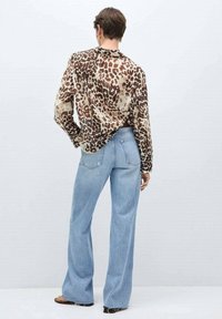 Personne vue de dos portant un chemisier marron à imprimé léopard et un jean à jambes larges bleu clair avec des sandales noires sur un fond uni.