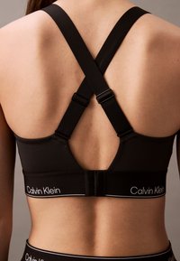 Vedere din spate a unei persoane care poartă un sutien negru Calvin Klein, cu bretele încrucișate reglabile și o bandă cu închidere cu cârlig și ochiuri.