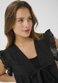 Blusa nera con maniche in pizzo arricciate, con scollo a V, davanti annodato e tessuto testurizzato. I dettagli delicati aggiungono interesse visivo.