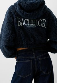 Navyblå kort fleecehuva med plyschtextur, med "BACHELOR OREGON STATE" broderat i vitt. Matchas med mörka jeans.