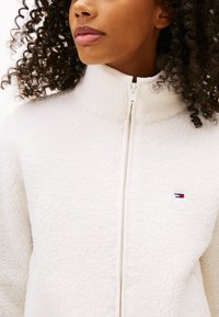 White fleece jacket with high collar and full zipper. Features a small embroidered logo in red, white, and blue on the chest. Soft texture.