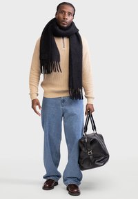 Beige stripete genser med et svart frynsete skjerf, løse blå jeans og brune sko. Holder en svart skinn duffelbag.