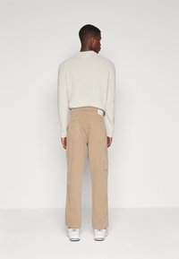 Beige Cargo-Hosen mit lockerer Passform, ausgestattet mit großen Seitentaschen und Gesäßtaschen. Kombiniert mit einem hellgrauen Strickpullover und weißen Sneakers.