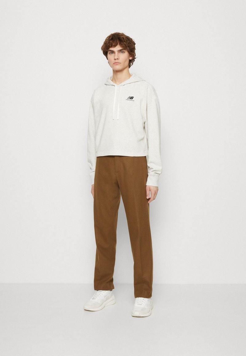 Sweat à capuche gris clair, doté d'un logo noir sur la poitrine ; associé à un pantalon en texture marron et des baskets blanches.