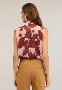 Donna che indossa una blusa senza maniche rosa con grande stampa floreale rossa e pantaloni marroni a vita alta, in piedi di spalle contro uno sfondo semplice.