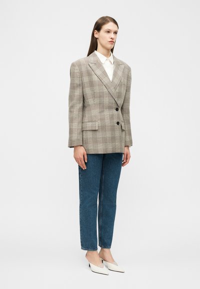 Frau trägt beigen karierten doppelt geknöpften Blazer, weißes Hemd, blaue gerade geschnittene Jeans und weiße Slingback-Pumps mit spitzer Schuhspitze, steht vor weißem Hintergrund.