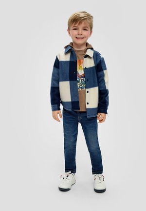 Marineblaue und cremefarbene Karo-Fleecejacke, geknöpft, mit einem braunen Grafik-T-Shirt darunter, kombiniert mit blauen Jeans und weißen Sneakers.