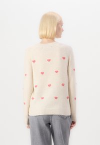 Pull crème avec une texture douce, arborant des motifs de cœurs roses éparpillés, des manches longues et un col et un ourlet côtelés.