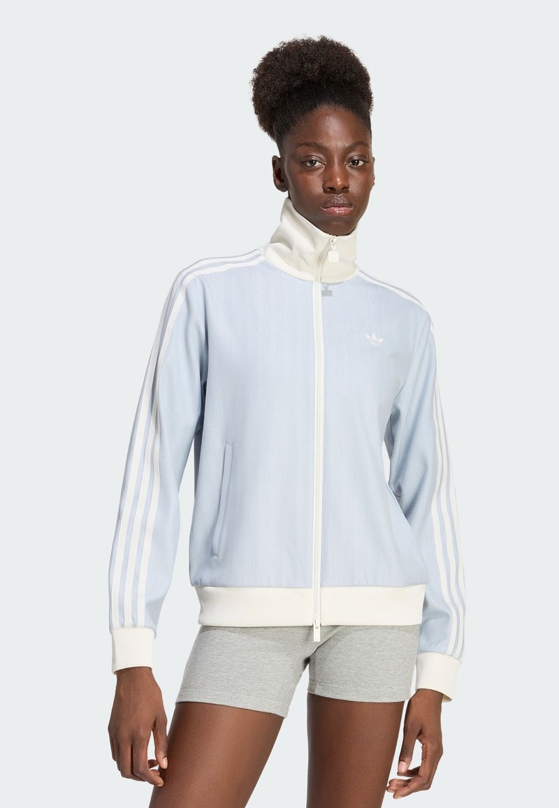 Femme portant une veste zippée bleu clair et blanche avec des rayures sur les manches et un short gris sur un fond uni.