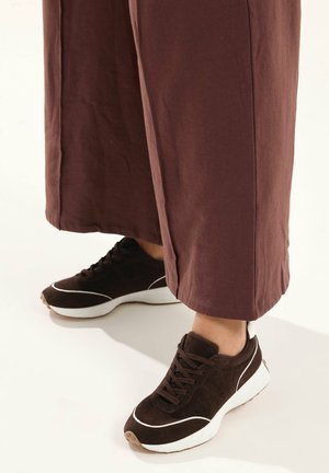 Mørkebrune sneakers med hvide detaljer båret med løse, vidde-ben brune bukser på en lys baggrund.