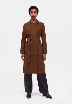 Un mannequin se tient devant un fond blanc, vêtu d’un manteau croisé marron ceinturé, d’un pantalon noir et de bottes à talons.