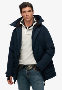 Veste isolante bleu marine avec une capuche amovible, dotée d'une fermeture éclair, de deux poches latérales et d'une doublure intérieure douce.