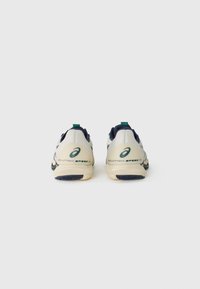 Lightweight tennis shoes in cream and navy, featuring a textured upper, a green accent loop, and a cushioned sole with patterned tread.