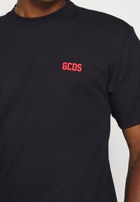 Černé tričko s krátkým rukávem a malým červeným logem "GCDS" na levé hrudi, které má na sobě osoba s tmavou pletí.