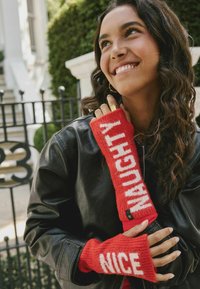 Guantes sin dedos de punto rojo que cuentan con el texto blanco "NAUGHTY" y "NICE" en las muñecas, con una textura suave y un diseño acanalado. Chaqueta de cuero negra debajo.