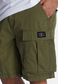 Pantalones cortos tipo cargo de color verde oliva, confeccionados en un tejido texturizado con un patrón de cuadrícula, que cuentan con bolsillos laterales y una etiqueta negra en el bolsillo derecho.