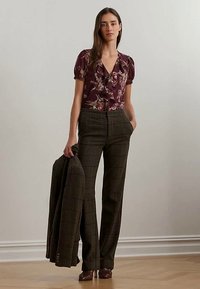 Kvinne iført en lilla blomsterbluse og brune rutete bukser, som holder en matchende brun rutete blazer i den ene hånden.