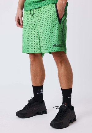 Man draagt groene korte broek met patroon, zwarte sokken en zwarte Nike sneakers, staat met één hand in de zak.