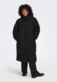 Pitkä musta toppatakki hupulla, jossa on tikattu kuviointi, etutaskut ja säädettävät kiristysnyörit kaula-aukossa. Tasainen pinta.