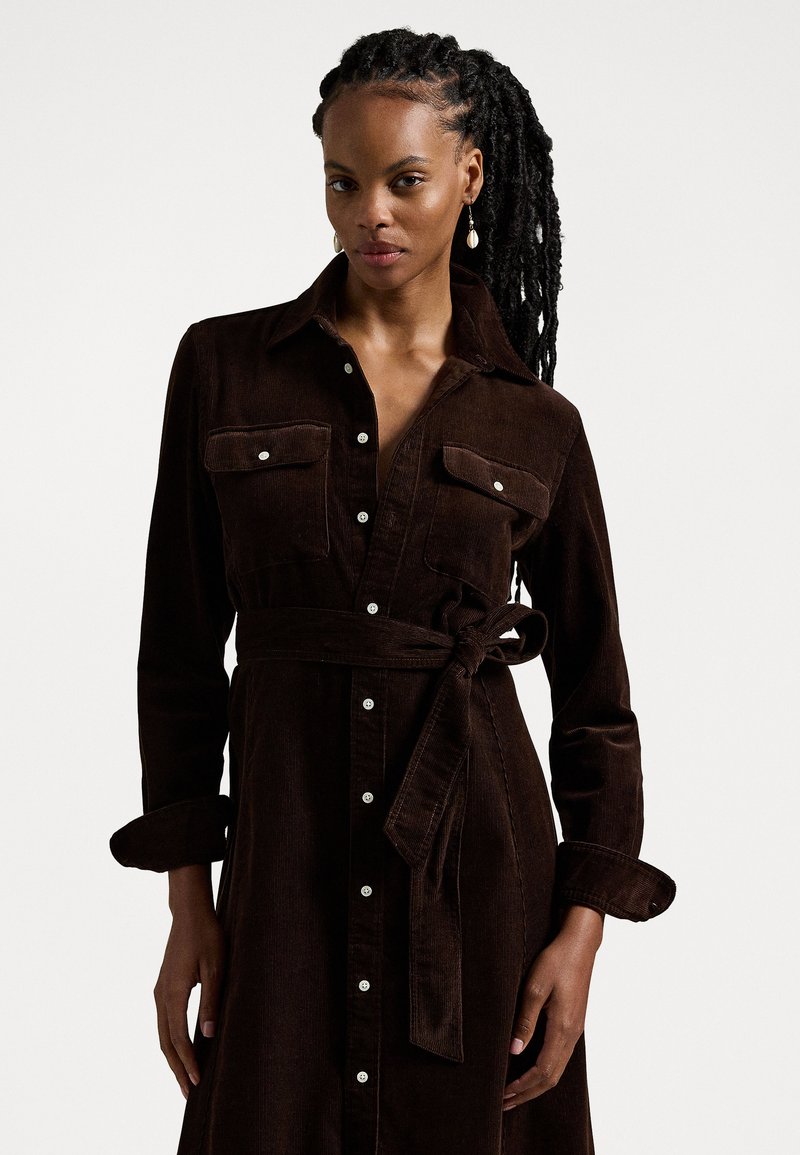 Brun fløjlskjole med krave, lange ærmer, brystlommer og taljebånd. Har knaplukning og en struktureret stof.