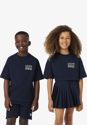 Dos niños sonrientes con conjuntos azul marino Lacoste Roland Garros Club, uno con pantalones cortos y camiseta, el otro con falda plisada y camiseta corta.