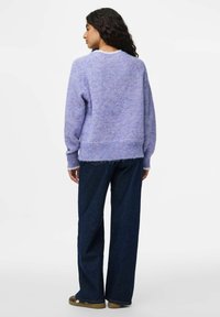 Femme avec de longs cheveux bouclés, portant un pull en laine de lavande, un pantalon large bleu foncé et des chaussures marron, de dos, sur fond blanc.