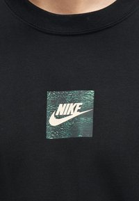 Μαύρο πουκάμισο με ένα μικρό πράσινο τετράγωνο που απεικονίζει το λευκό λογότυπο της Nike και το swoosh στο στήθος κάτω από το λαιμό.