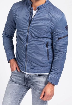 Mężczyzna noszący dopasowaną niebieską kurtkę bomber z pikowanymi detalami, kieszeniami na suwak i jasnymi dżinsami, ręka w kieszeni.