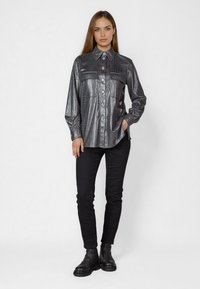 Metallisch dunkelgraues Hemd mit Krokodilstruktur, drei Knöpfen auf der Vorderseite und zwei Brusttaschen. Kombiniert mit schwarzen Slim-Fit-Hosen und Stiefeln.