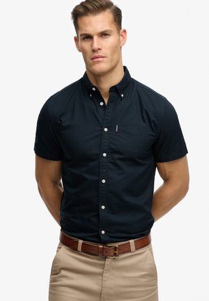 Hombre vistiendo una camisa azul marino ajustada de manga corta con botones y bolsillo en el pecho, y pantalones color caqui, de pie con las manos detrás de la espalda contra un fondo blanco.