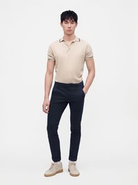 Jeune homme debout portant un polo beige à manches courtes, un pantalon bleu marine roulotté, des chaussettes blanches et des baskets beiges sur fond blanc uni.