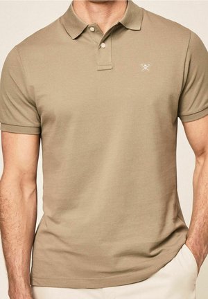 Mand iført en beige kortærmet polo t-shirt med to knapper og et lille hvidt broderet logo på brystet.