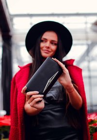 Black leather wallet with a glossy silver accent, rectangular shape, held in a hand, against a blurred red coat background.