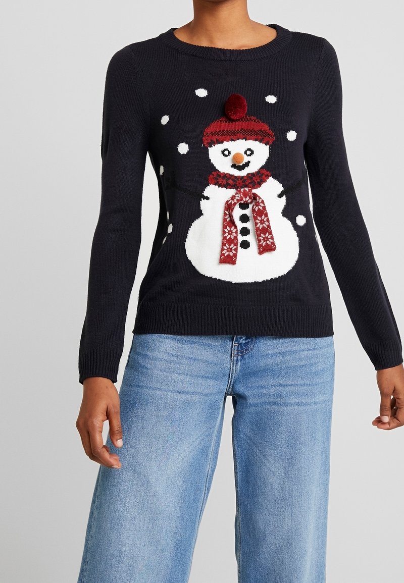 Persona che indossa un maglione nero con un pupazzo di neve bianco, cappello e sciarpa rossi, e puntini di neve che cadono, abbinato a jeans azzurri chiari.