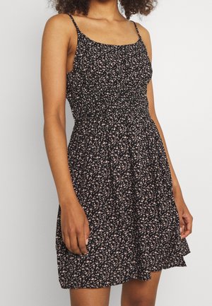 Kvinde iført en ærmeløs sort kjole med små pink og hvide blomsterprint og talje med rynkede detaljer, stående mod en ensfarvet baggrund.