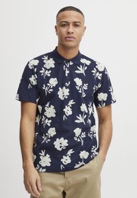 Marineblå polo skjorte med stort hvitt blomster mønster, korte ermer, to-knapps placket og subtil tekstur. Kombinert med lysfargede bukser.