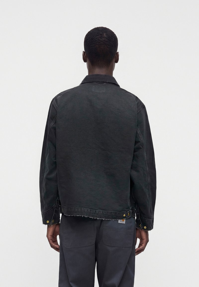 Mann von hinten gesehen, der eine schwarze Jeansjacke und dunkelgraue Hose vor einem schlichten hellen Hintergrund trägt.