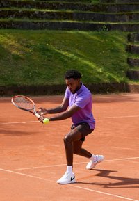 Tennisspelare i en lila polo och svarta shorts, svingar ett racket för att träffa en gul tennisboll på en grusplan. Grön gräs i bakgrunden.